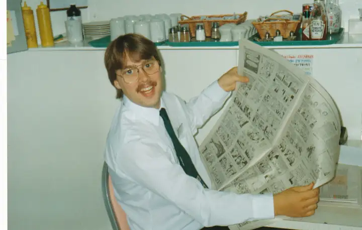 Jim Boykin in a white shirt and tie, sitting with a big newspaper comics page open and a wide grin